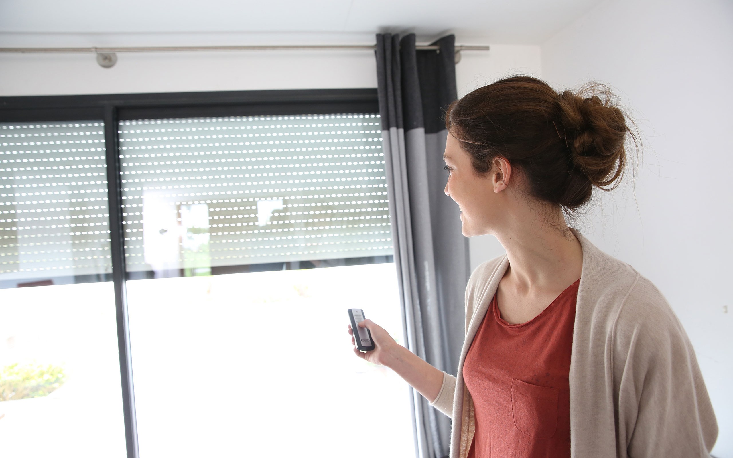 a smiling woman in a red shirt trying a motorized blind at home with remote control